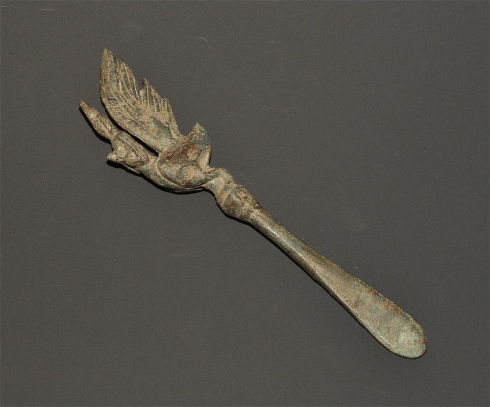 Cambodian Bronze Winged Figure Cosmetic Applicator: An undated archaistic cast applicator for cosmetics and unguents formed as a rounded blade with balustered top surmounted by a figurine formed as a cockerel with the head replaced by a Buddha figure w