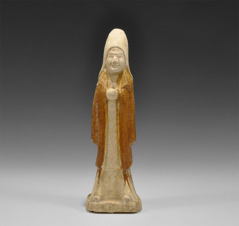 Chinese Glazed Ceramic Soldier Figurine: Tang Dynasty, 618-906 AD A figurine of a standing soldier in tall hood and long coat over a floor-length robe, standing on a square base; the figure with hands drawn up to the chest, pierced; the coat