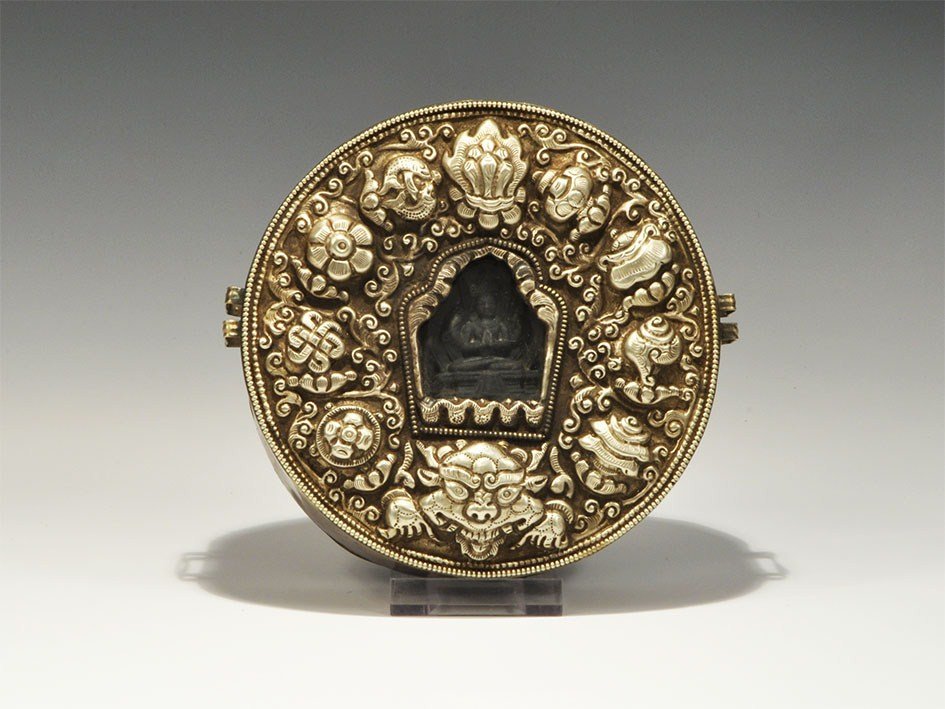 Tibetan Buddhist Shrine Mount in Silvered Display: Circa 19th century AD A carved stone mount in the form of the Buddha seated on a low throne visible through a glass pane in the lid; the drum copper with two pairs of strap-sliders; the lid silvered w
