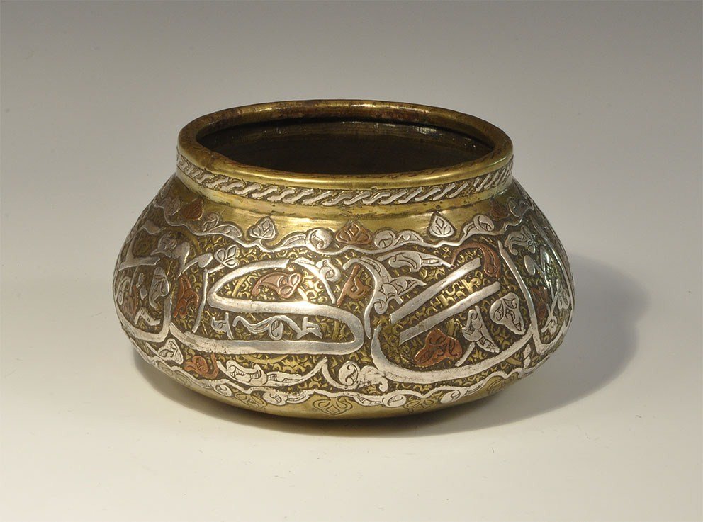 Islamic Bronze Epigraphic Squat Bowl: 19th century AD A squat bronze vessel with rolled rim inlaid with a silver guilloche; the body textred with inlaid silver vineleaf design, displaying reserved calligraphic text in silver with copper a