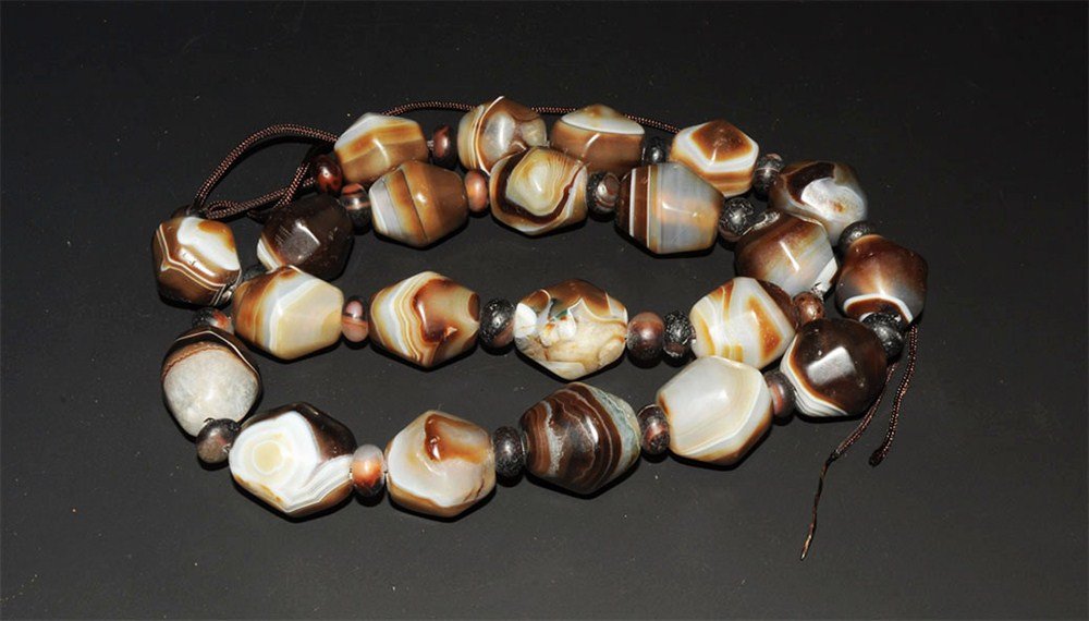 Near Eastern Bactrian Agate Polyhedral Beads Group: An undated restrung group of graduated banded agate beads comprising twenty-two large polyhedral beads and twenty-one globular seed bead spacers. 252 grams, 62 cm (24 1/2"). Property of a London Gentl