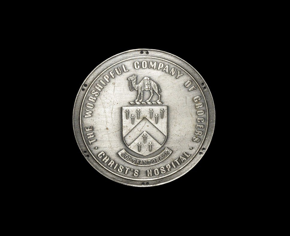 Vintage Worshipful Company of Grocers Badge: 20th century AD. A large, unifacial silvered metal badge with six pairs of sewing holes around edge depicting the Grocer's Arms surmounted by a camel and with 'God Grant Grace' motto below, with THE W