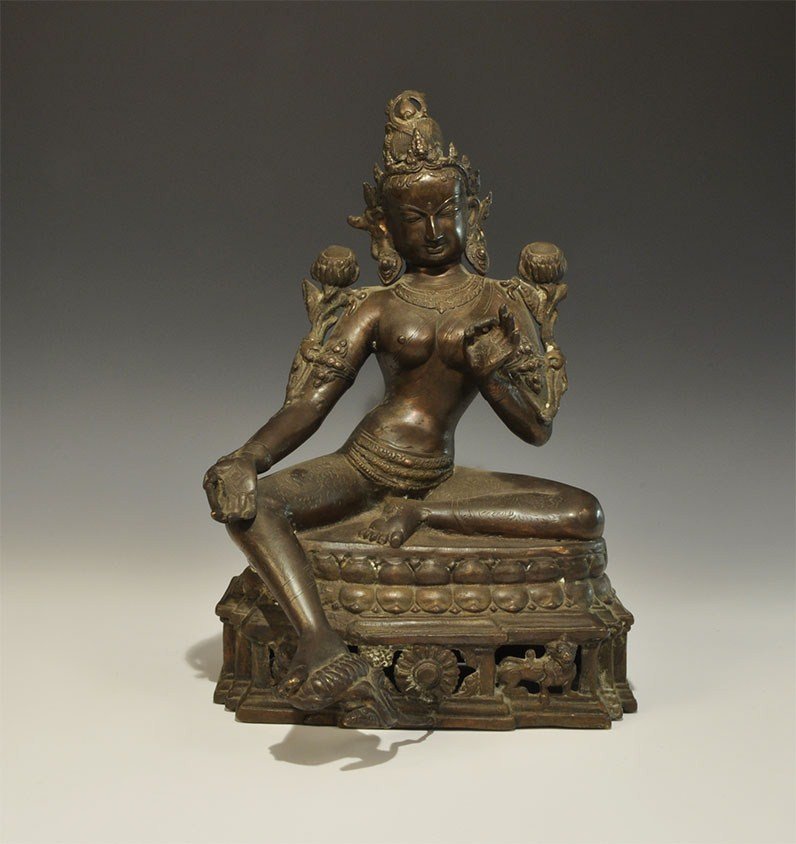 Thai Bronze Goddess Figurine: 19th century AD. A hollow-cast figurine depicting a goddess seated on a low bench, wearing a tall headdress, arm-rings, necklace and loincloth; the left hand raised in the Buddhist gesture of teaching