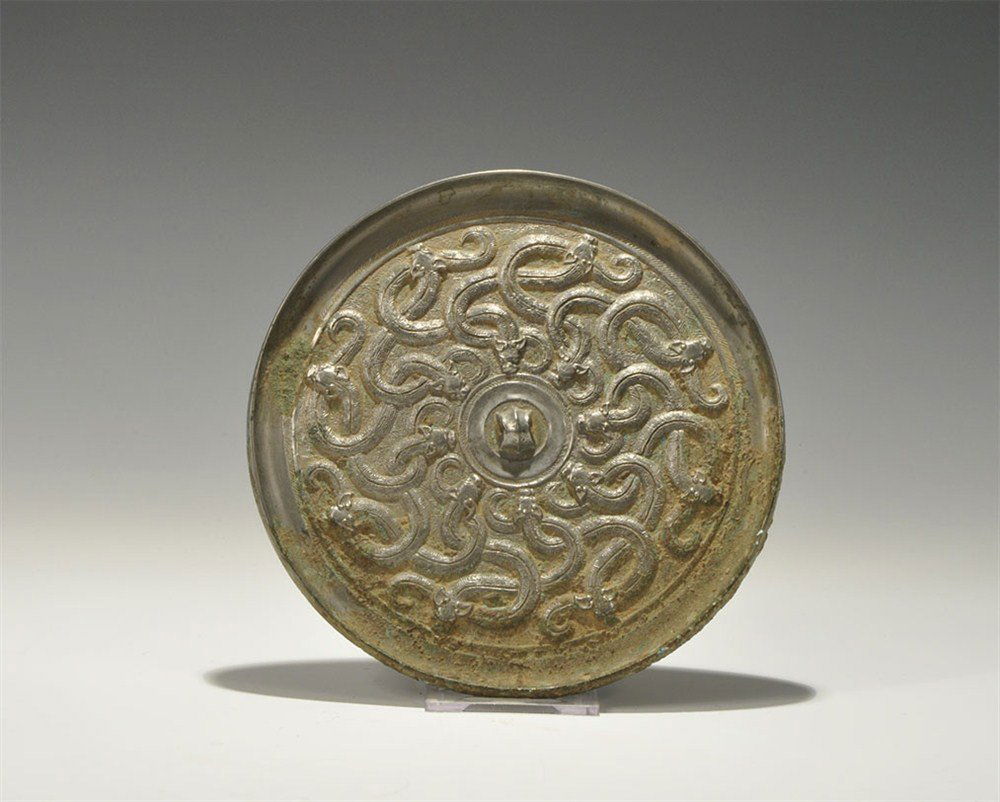 Chinese Style Tinned Bronze Interlaced Serpents Mirror: An undated archaistic cast-bronze disc mirror with one tinned and polished surface; to the reverse, a central pierced ribbed lug surrounded by interlaced s-shaped serpents, eight facing inwards and ei