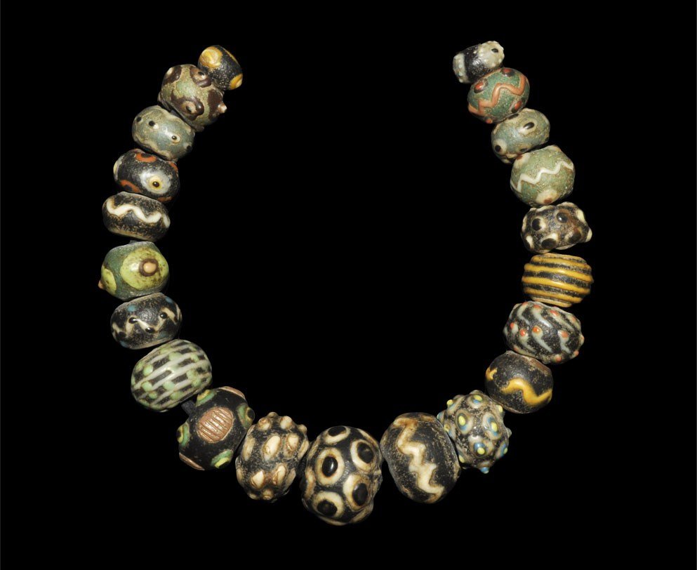 Roman Syrian Style Glass Bead Group: An undated restrung necklace comprising twenty-one large graduated beads; various colours, some with eye detailing, applied trails in zigzags or bands. 335 grams, 44 cm (17"). London collection; acqui