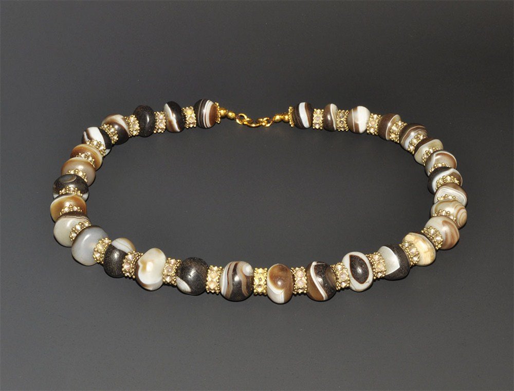 Near Eastern Bactrian Style Banded Agate Bead Necklace: An undated restrung necklace comprising thirty-three oblate banded agate 'Suleimani' beads, with spacers comprising gold seed beads, gold disc rosettes and agate disc beads; modern clasp. 178 grams, 5