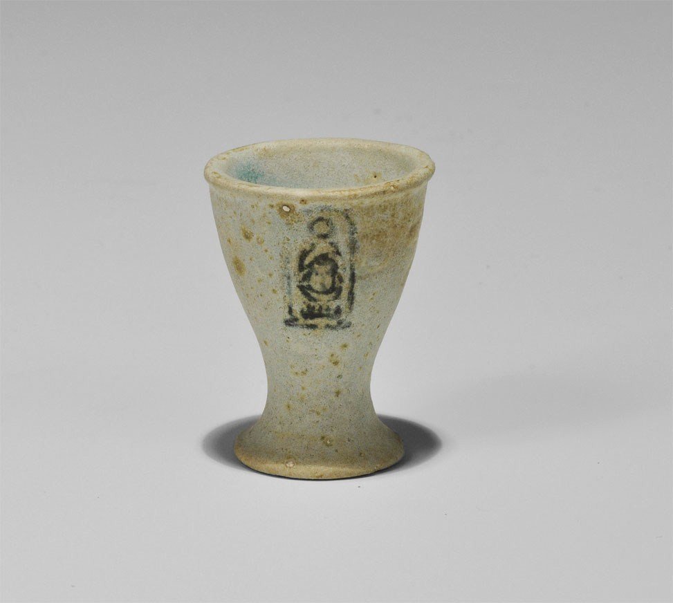 Egyptian Offering Cup with Cartouche of Thuthmosis III: Circa 1450 BC. A pale blue faience miniature cup with flat base, waisted with rolled rim; below the rim a dark blue cartouche containing the name of the pharaoh Tuthmosis III in hieroglyphic text. 18