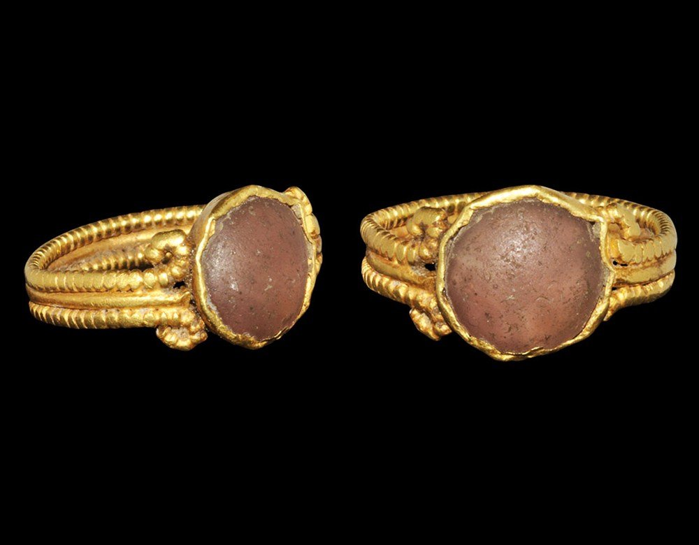 Roman Gold Cabochon Ring: 2nd-3rd century AD. A gold finger ring, the hoop formed from three parallel rods, the outer two pelletted and ends scrolled; the bezel a discoid panel with inset pale glass cabochon. 4.91 grams, 22 mm