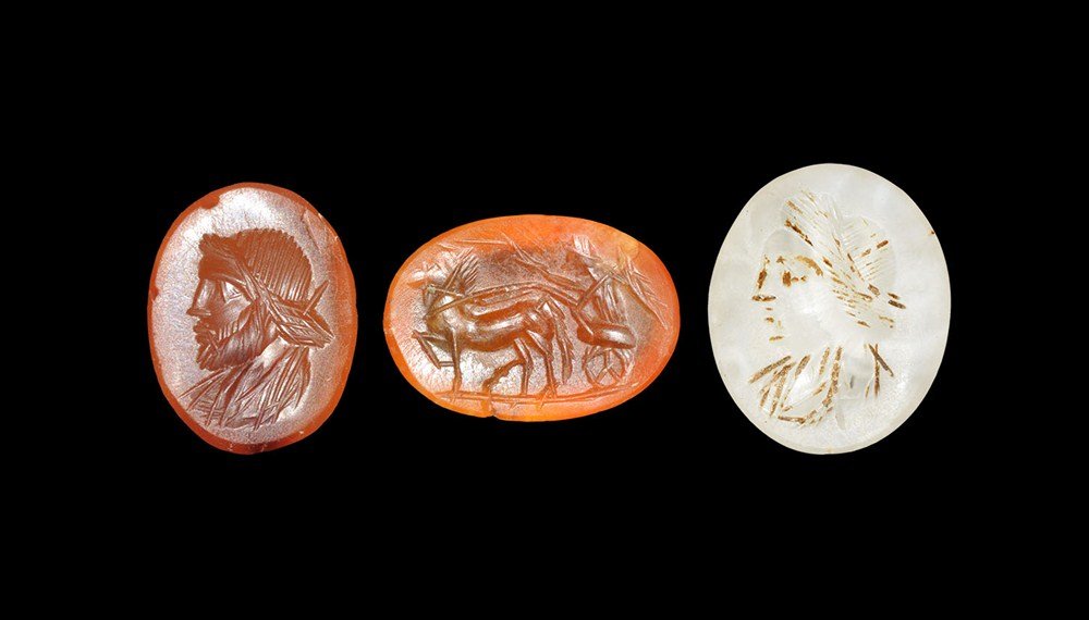 Roman Stone Chariot Motif Intaglio: 1st-3rd century AD. An orange hardstone intaglio with chariot motif, offered with two possibly 19th century intaglios: one with bearded male bust, the other in white hardstone with female bust in prof
