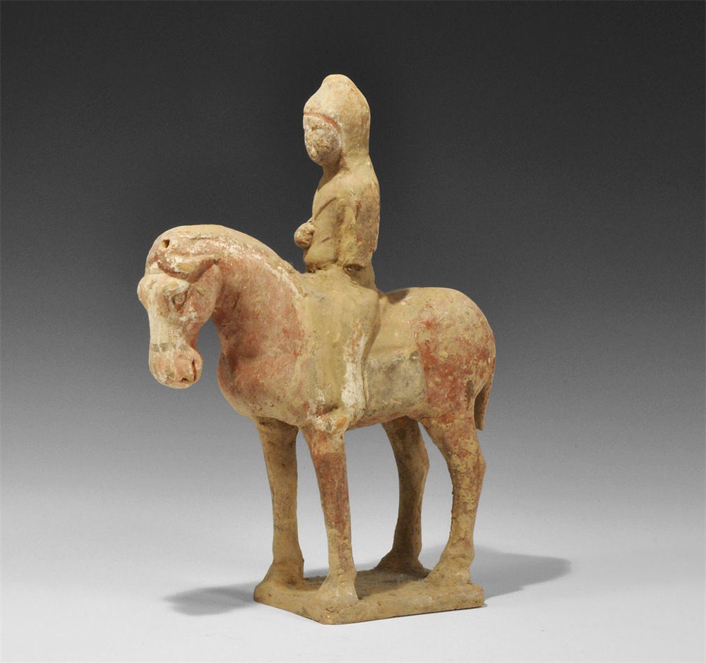Chinese Horse and Rider Figurine: Tang Dynasty, 618-906 AD. A ceramic figurine of a standing horse and rider; the horse with head bowed, on a square base; the rider in hood and riding coat, hand drawn up to the chest, pierced; painted