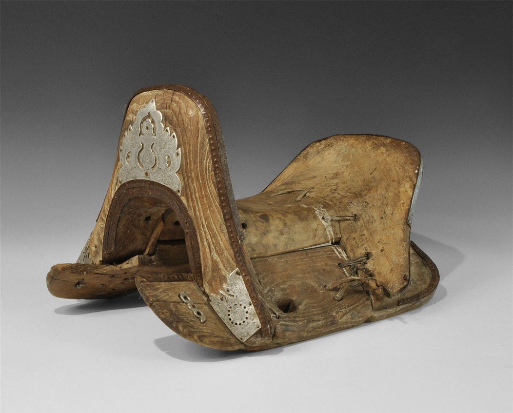 Chinese Tibetan Wooden Saddle with Silver Appliques: 18th-19th century AD. A carved wooden saddle fabricated from five components laced together with thong: head and cantle with iron strips and silver appliques fore and aft; two lateral shaped boards wi