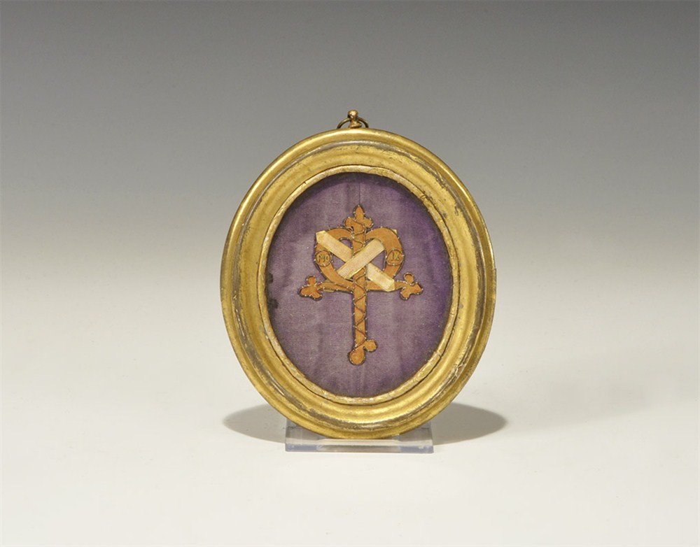 Christian Pius IX Reliquary Plaque: A reliquary plaque displaying an embroidered intertwined crosses-and-heart motif inscribed 'PIO IX' from the vestments of Pope Pius IX (1792-1878); mounted on velvet in an oval gilt wood glazed frame