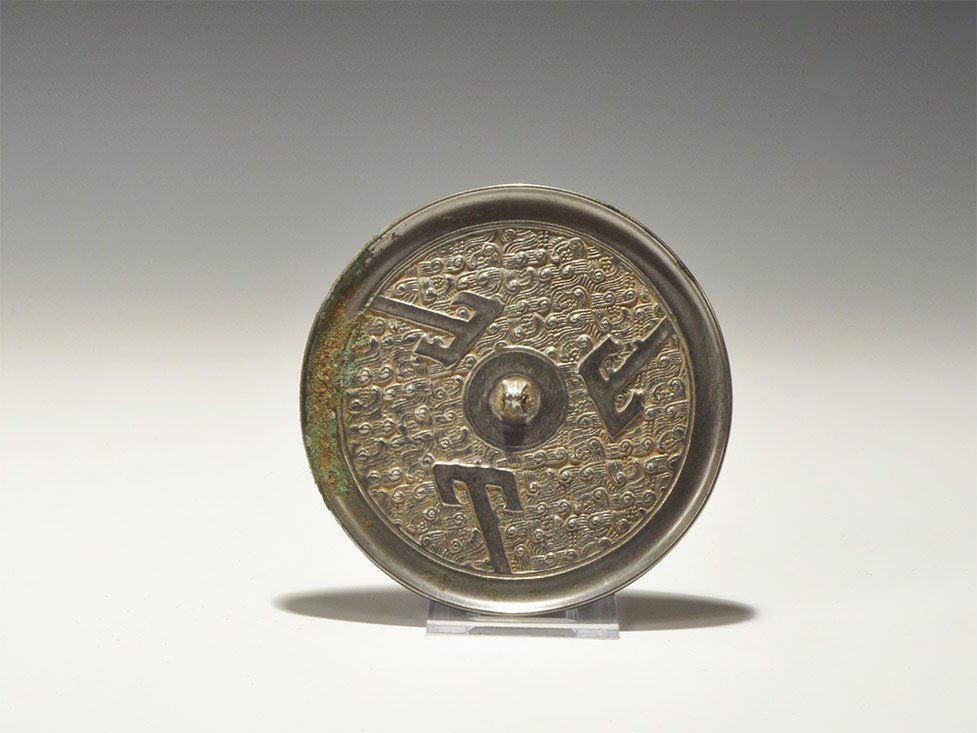 Chinese Style Bronze Mirror with Field of Scrolls: An undated archaistic discoid bronze mirror with one tinned surface, the reverse with central pierced lug on a field of scrolls, three T-shaped elements emanating from the plain border. 241 grams, 12.