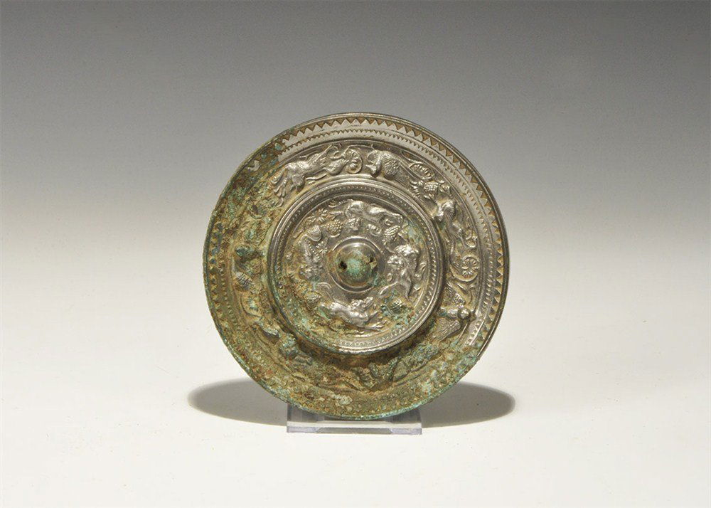 Chinese Style Bronze Figural Mirror with Birds, Fruit a: An undated archaistic discoid bronze mirror with one tinned surface, the reverse with central pierced lug surrounded by four running lions amid fruit, outside this a band of birds, fruit and animals.