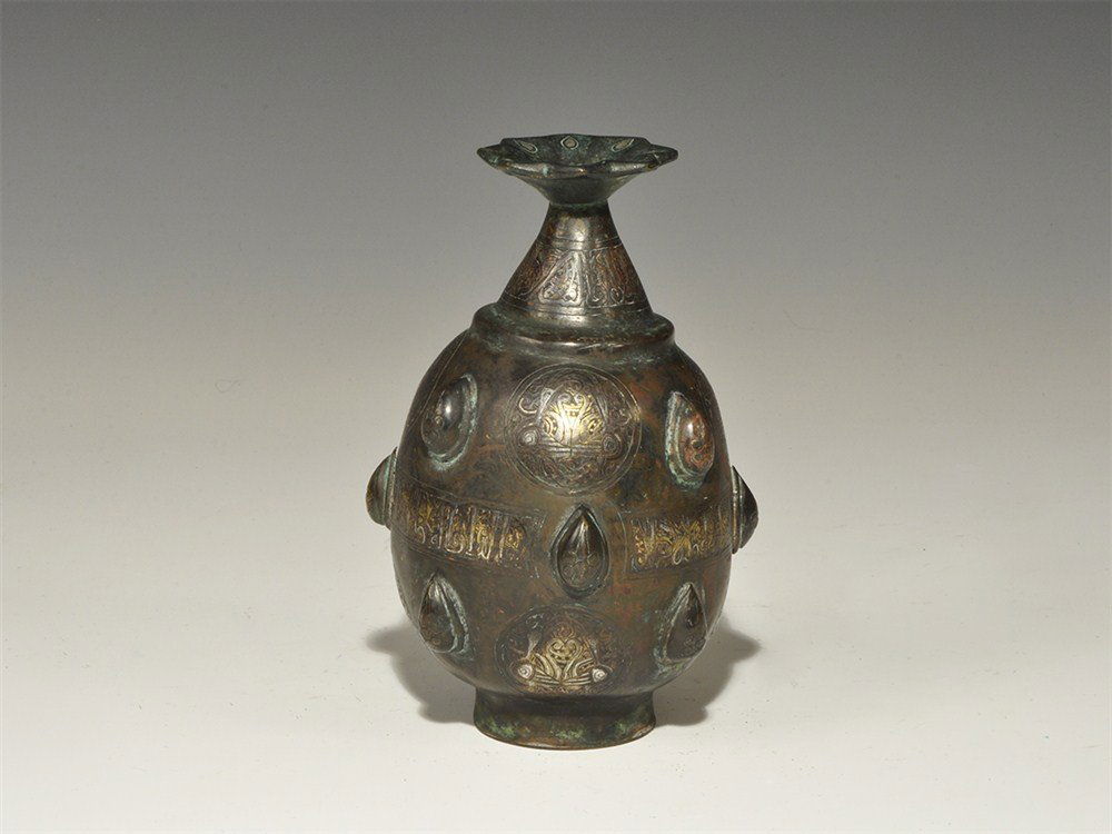 Islamic Style Bronze Silver-Inlaid Calligraphic Perfume: An undated archaistic piriform bottle with flat base and conical neck, flared octofoil rim; the body with raised ovoid bosses; bands of silver-inlaid calligraphic text to the neck and centre, figural