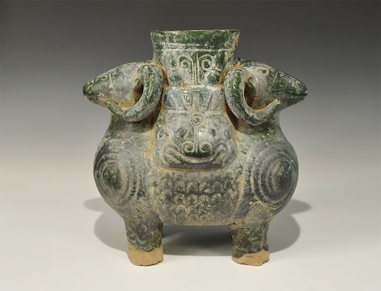 Chinese Style Ceramic Ram Vessel: An undated archaistic green-glazed vessel in the form of two addorsed rams with textured detailing to the horns and fleece; the central portion to each side a grotesque horned face with tubular neck a