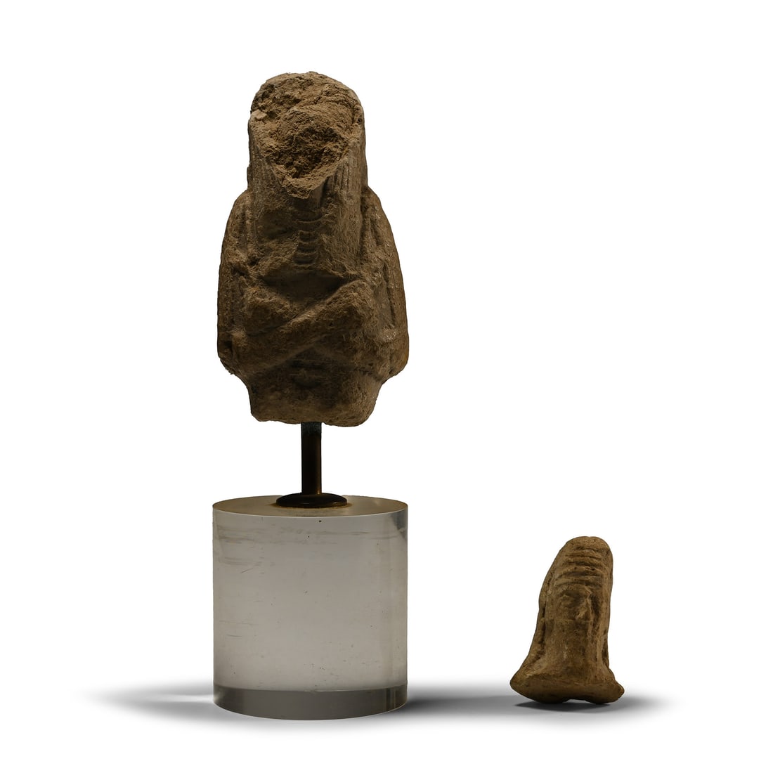 Babylonian Head and Egyptian Shabti Group: Mid 1st millennium B.C. and later Comprising of a male head from a figurine, and a possible Grand Tour fragment of a shabti with tripartite wig and arms crossed at the chest, mounted on a custom-made