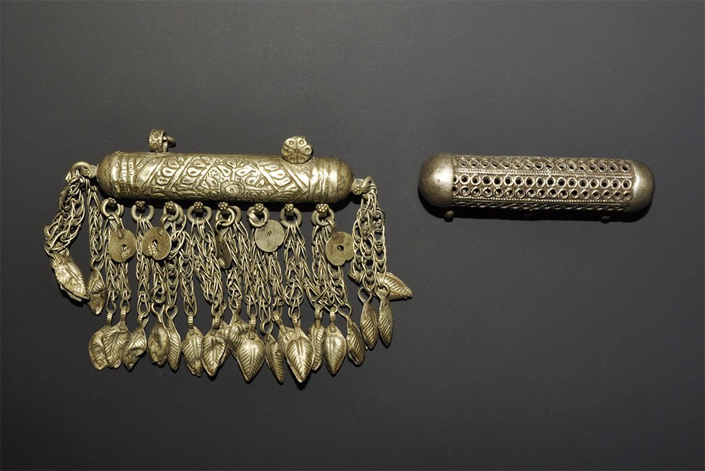 Islamic White Metal Scroll Holder Pendant Group: A group of two undated archaistic white metal pendants comprising: one tubular with domed cap ends, three suspension loops, openwork wall; one similar with domed ends, scale pattern to one face and fo
