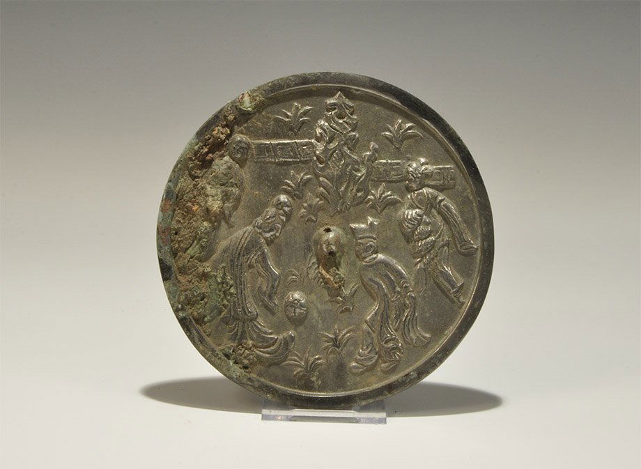 Chinese Style Bronze Figural Mirror with Ball-Players: An undated archaistic discoid bronze mirror with one tinned surface, the reverse with central pierced lug surrounded by a scene of figures in loose robes amid vegetation playing with a ball. Property