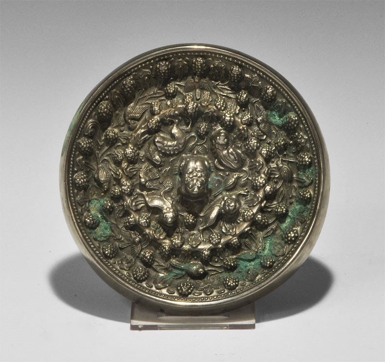 Chinese Tinned Bronze Mirror with Fruit, Monkeys and Bi: Tang Dynasty, 618-906 AD. A small cast-bronze rosette disc mirror with one tinned and polished surface; to the reverse, a central pierced zoomorphic lug within concentric bands of scrolled foliage wit