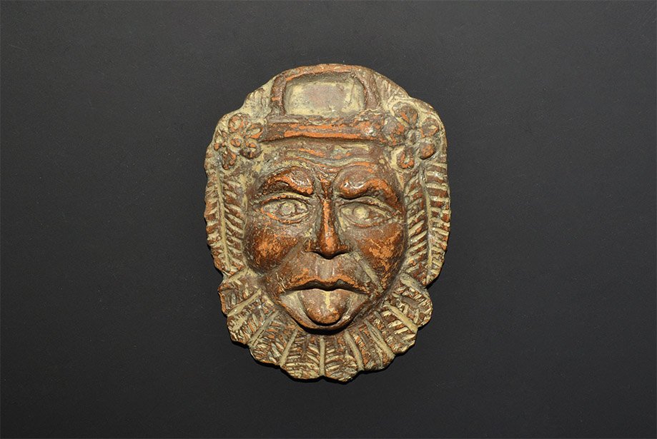 Roman Ceramic Dionysus Mask: 1st-2nd century AD. A small convex terracotta mask of the god Dionysus with flowers at the temples, leaves to the sides of the face and beard, the eyes wide open and tongue protruding; rectangular rec