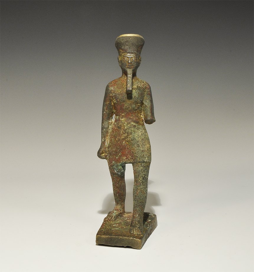 Egyptian Style Bronze Figurine: An undated archaistic cast figurine on a trapezoidal base depicting a barefoot figure in a short kilt and tall hat, with plaited beard and collar to the shoulders; hollow to the underside. Property of