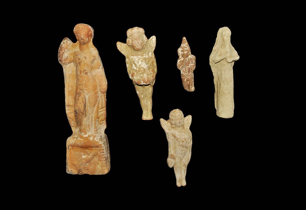 Greek and Roman Figure Group: 4th century BC-2nd century AD. A mixed group of ceramic figures of various dates comprising: a hollow-cast Tanagra figurine of a loosely-draped female on a rectangular base; a winged putto holding bun