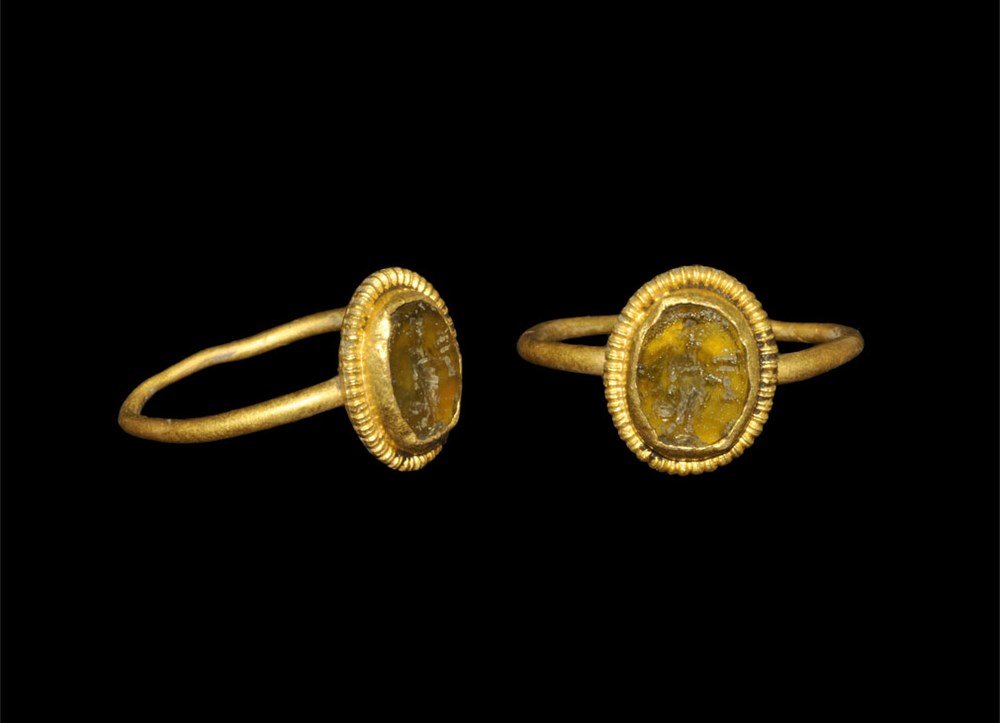 Roman Gold Finger Ring with Fortuna Intaglio: 1st-2nd century AD. A round-section hoop with elliptical bezel, filigree border and central brown glass cloison intaglio of Fortuna standing with one hand extended. 1.86 grams, 18 mm overall, 14.92 mm