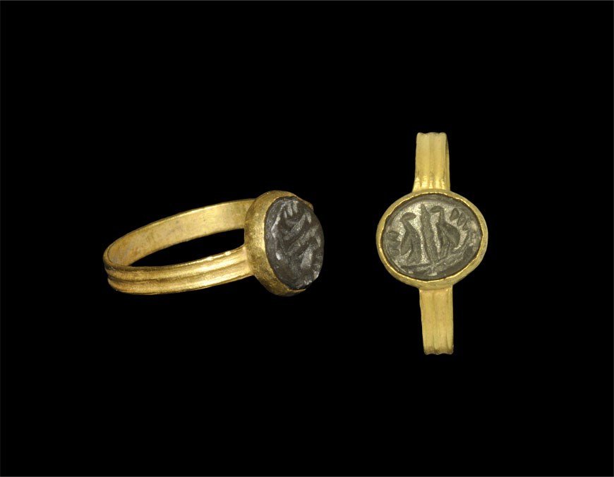 Roman Gold Finger Ring with Two Birds Intaglio: 2nd-3rd century AD. A flat-section reeded hoop and elliptical bezel with chamfered edge enclosing a brown hardstone intaglio of two birds facing an upright post on a baseline. 3.05 grams, 20 mm overal