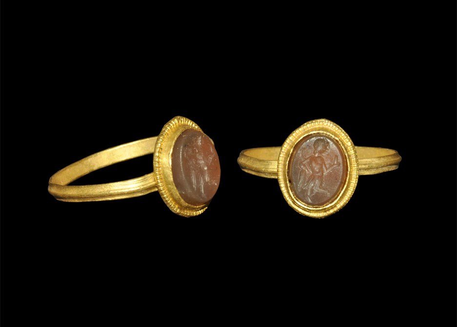 Roman Gold Finger Ring with Standing Figure Intaglio: 2nd-3rd century AD. A T-section hoop and elliptical bezel with filigree rim supporting a carnelian intaglio depicting a standing figure holding a rod in the crook of the right arm. 2.12 grams, 21 mm o