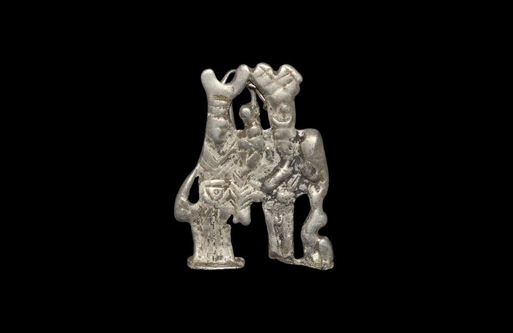 Roman Silver Harpocrates and Isis Plaque Amulet: 1st century AD. A cast silver amuletic plaque in the form of the god Harpocrates with finger raised to the mouth and his mother Isis to his right; two attachment loops to the reverse. 10 grams, 39 mm 