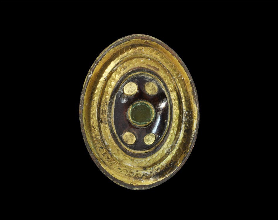 Roman Gilt-Bronze Umbonate Brooch with Later Inlays: 2nd century AD. An elliptical umbonate plate brooch with central raised cell, two outer bands of guilloche and chevron motifs; the cell set with a cabochon garnet; later modified with four gold studs