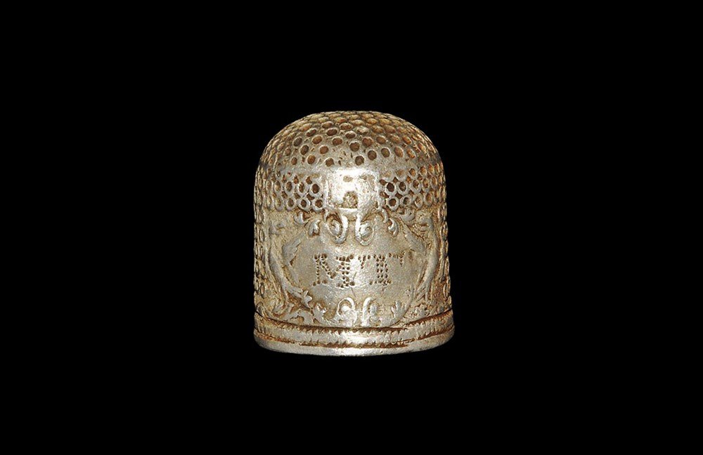 Post Medieval Silver Thimble with Initials: Circa 1650 AD. A delicate domed thimble with roundel texturing to the sidewall and top; a large rococo panel to the sidewall with pseudo-heraldic supporters and mural crown above, the initials 'MT' ex