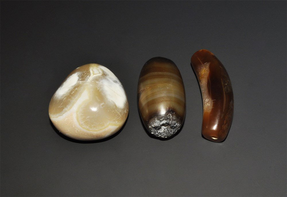 Middle Eastern Agate Bead and Polished Pebble Group: A mixed group of highly polished undated archaistic beads comprising: a russet -coloured Elbow bead, a natural barrel-shaped banded agate bead, and a pale agate pebble. 316 grams total, 57-69 cm (2 1/
