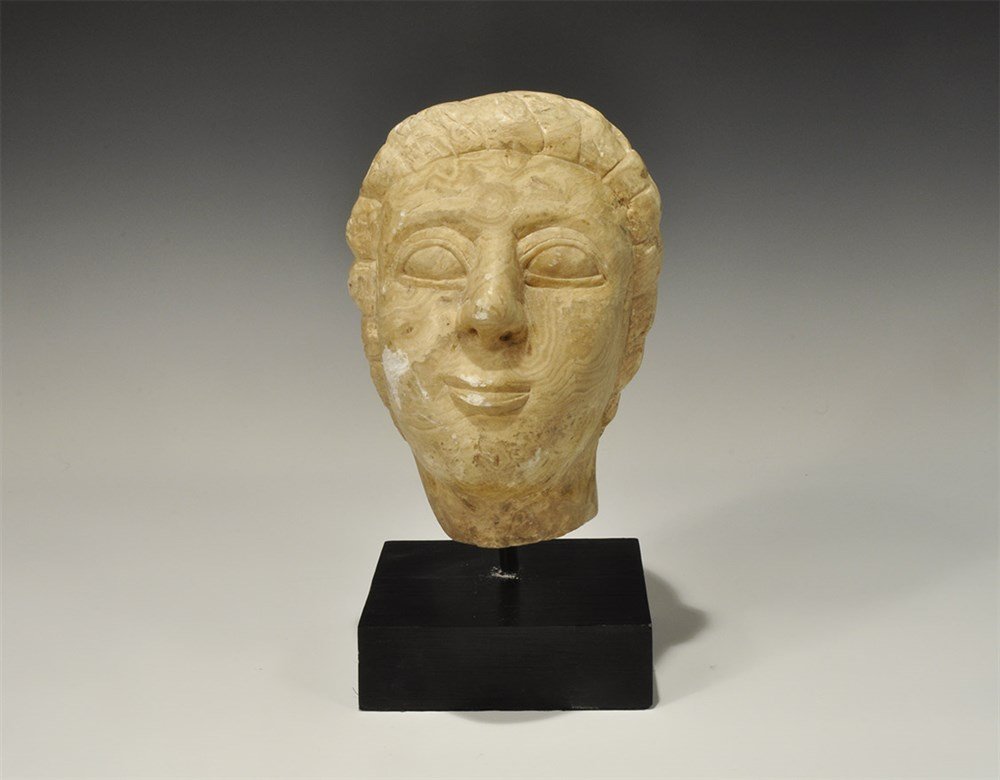 Near Eastern Style Alabaster Head: An undated archaistic carved female head with fleshy features and dressed hair; one a custom-made display stand. 7 kg, 30.5 cm (12"). Property of a London Gentleman; acquired before 2000.