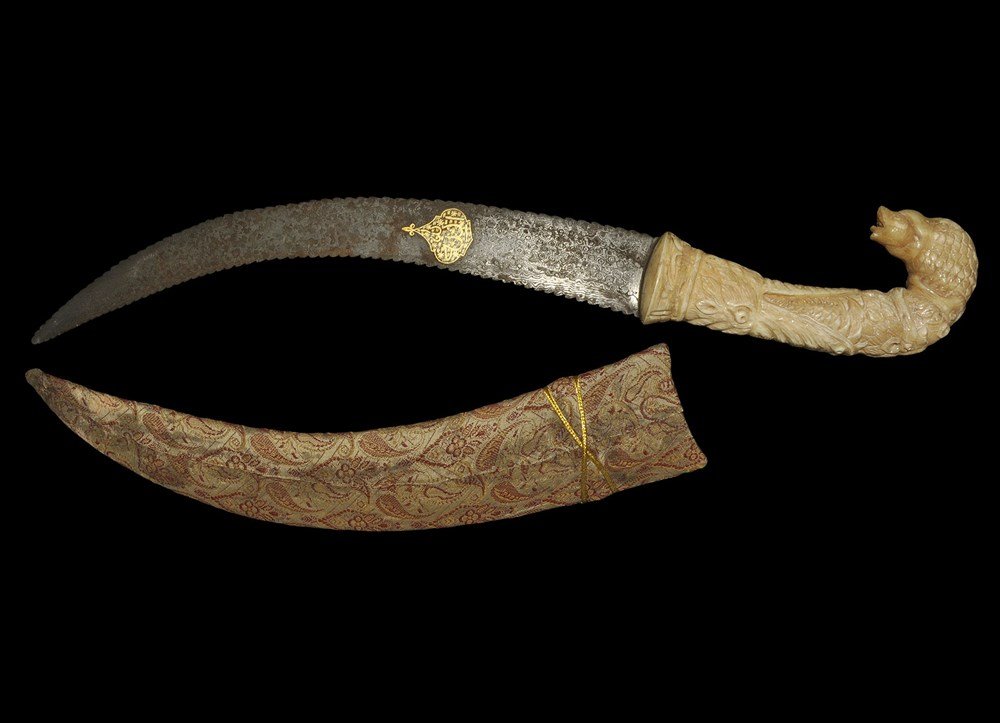 Indian Style Dagger and Scabbard: An undated archaistic dagger with curved blade and zoomorphic carved composition handle; the blade with serrated edge, inlaid motif to one face, unsharpened; the handle carved a a wolf's head with an 