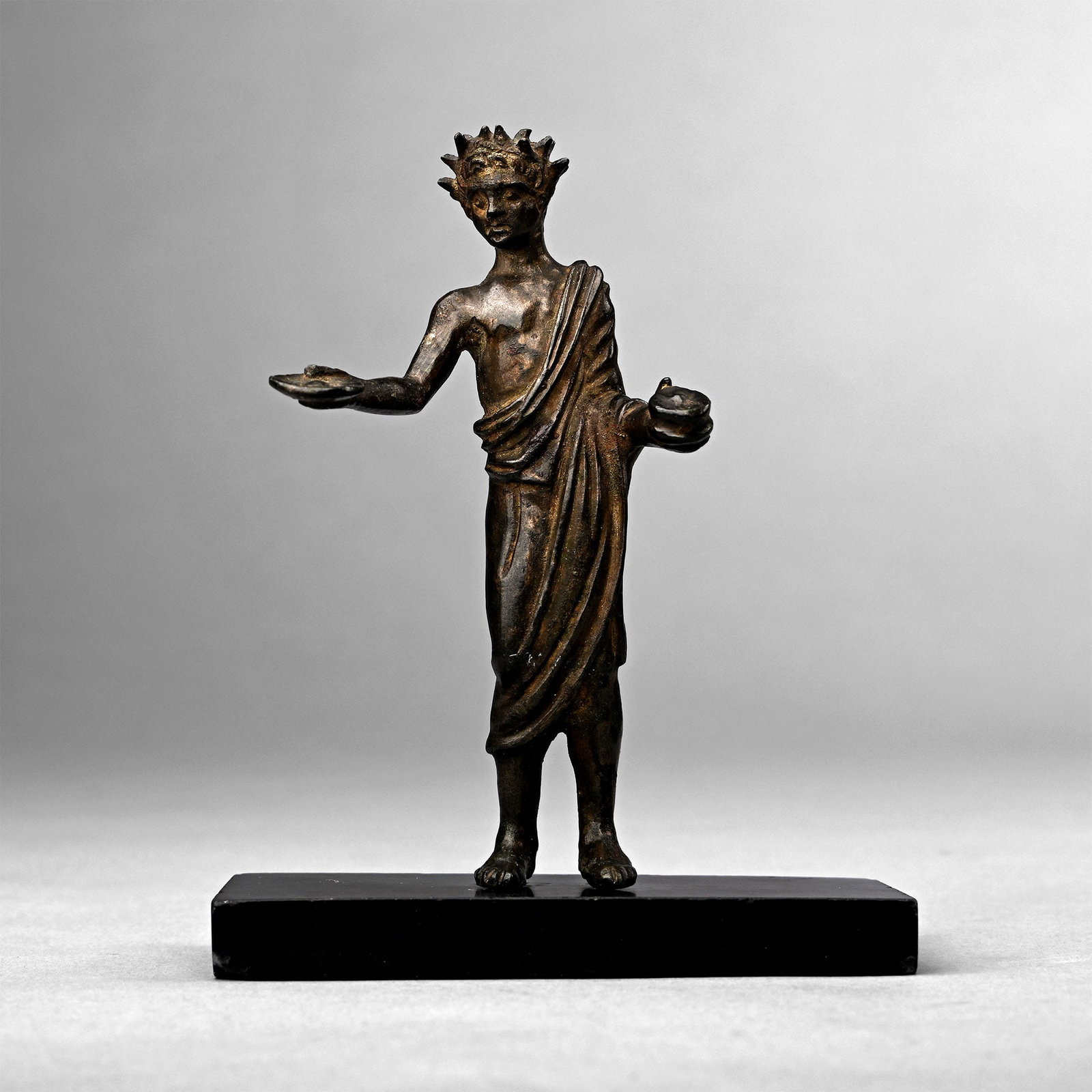 Etruscan Bronze Votive Figure of Genius Sacrificulus: 6th - 5th century B.C. Depicting a ‘genius’ making a sacrifice, his head surrounded by a radiate crown or a wreath of laurel leaves arranged in rays; bare-chested with a cloak (tebenna) falling ov