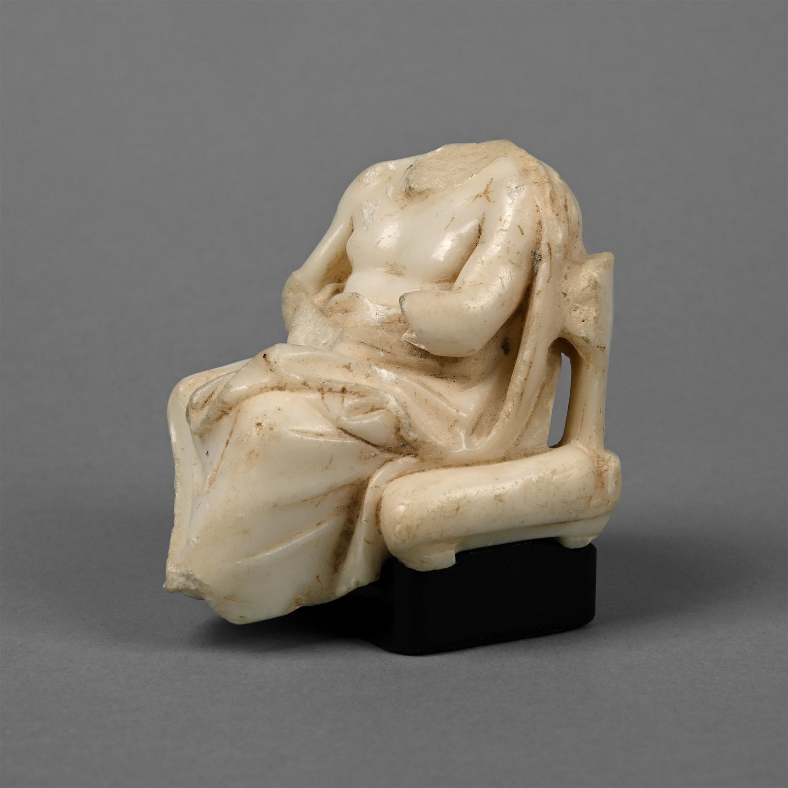 Greek Marble Figure of a Seated God or Philosopher: Hellenistic, 3rd-1st century B.C. Carved in the round male figure in a seated pose wearing a loosely draped pallium on the shoulders and lower body, legs crossed at the knees; openwork frame to the ch