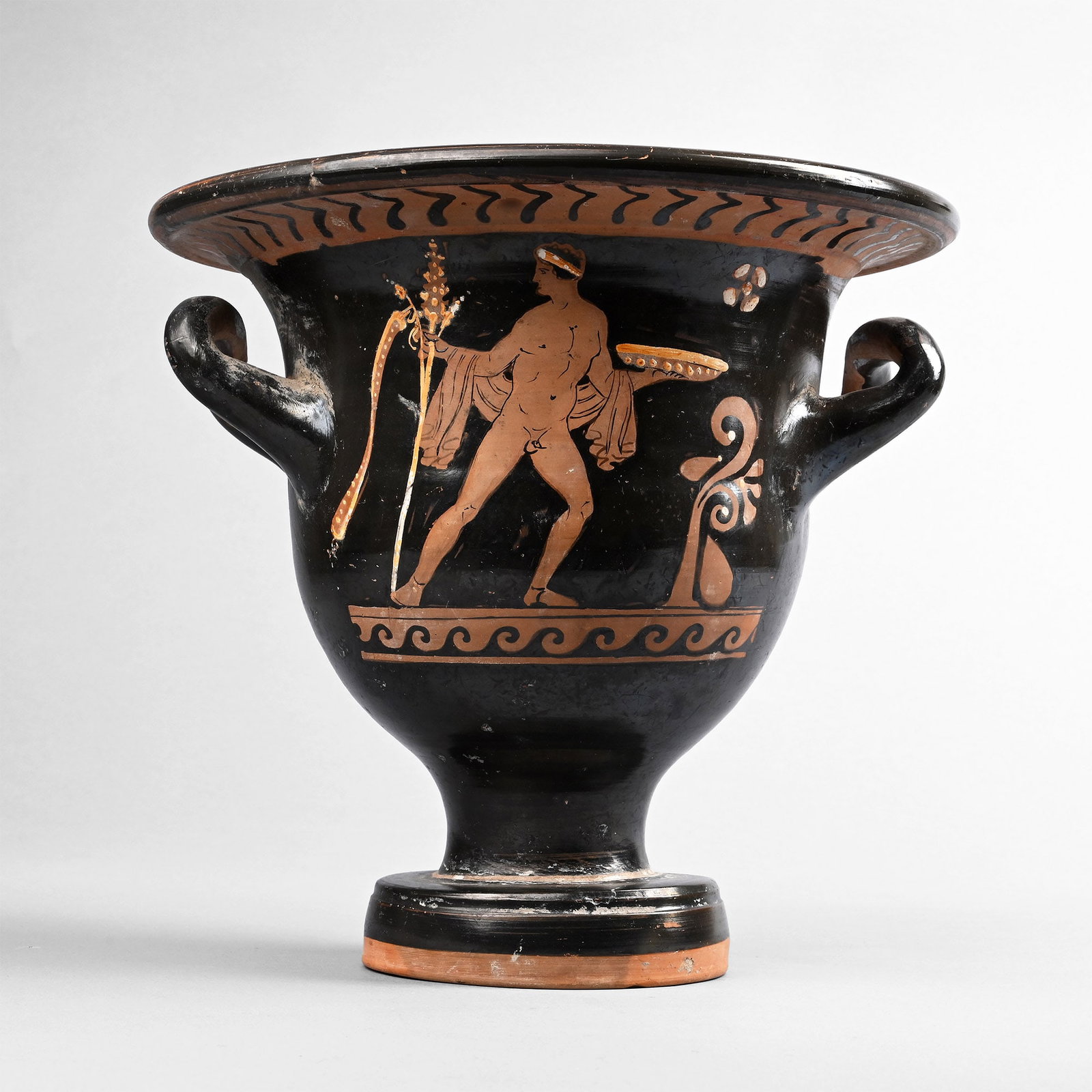 Greek Red-Figure Bell Krater Belonging to the Chevron Group: Apulia, 4th century B.C. With reserved herringbone band beneath the rim, wave pattern to be the baseline; Side A: young man with thyrsus and pendant streamer holding a phiale, a loose mantle draped fr