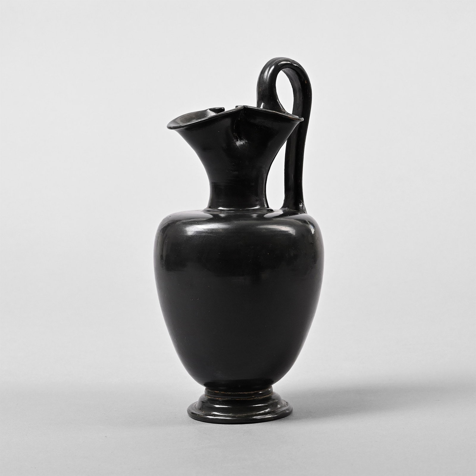 Large Greek Black-Glazed Trefoil-Lipped Oinochoe: Apulia, 5th century B.C. With a trefoil mouth, an applied D-section handle and a tiered foot. Cf. The Metropolitan Museum, accession numbers 81.10.337 and 17.194.743, for similar in glass; Beazley, J.