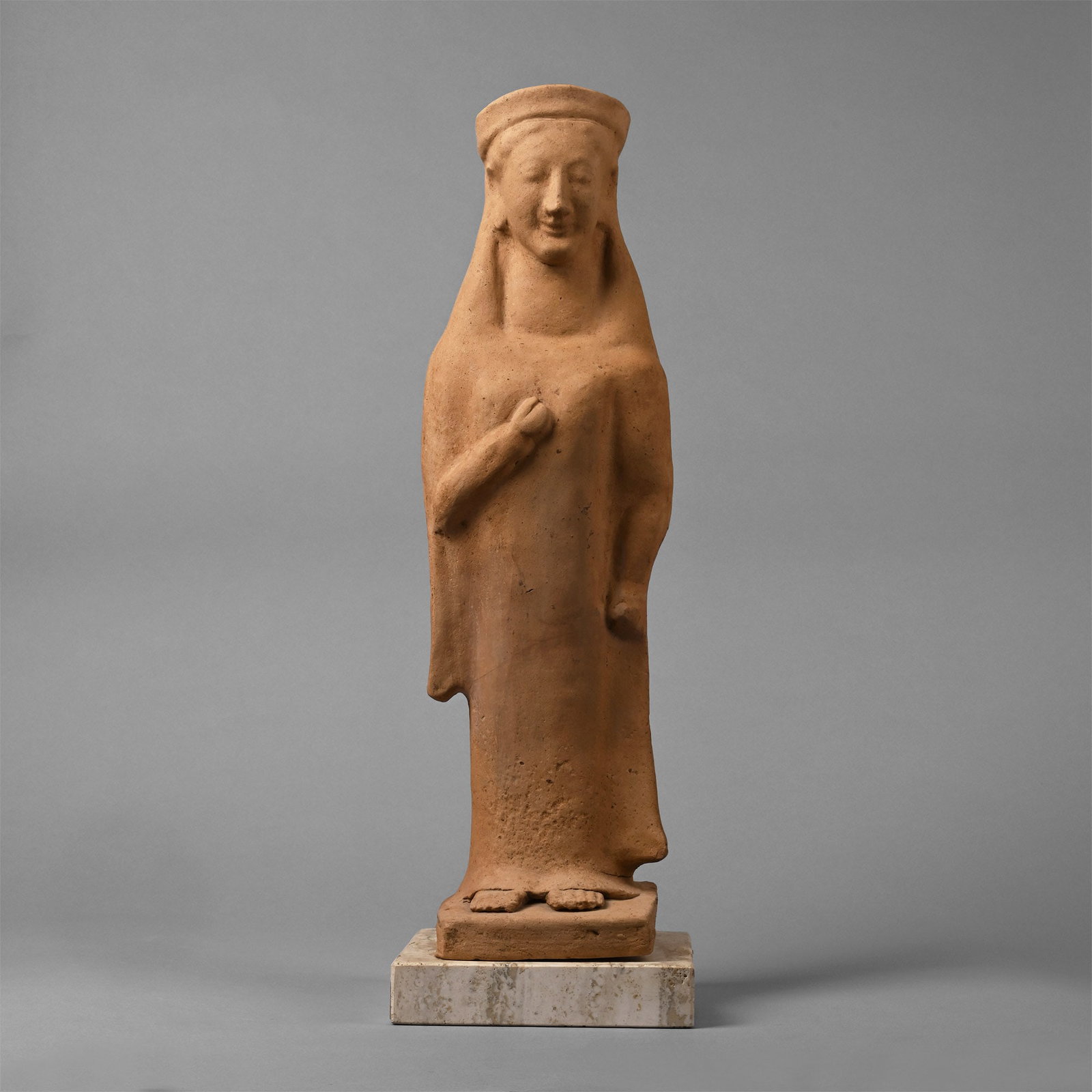 Large Greek Terracotta Figure of a Kore: 6th century B.C. Hollow-formed with scaphoid headdress, long hair hanging behind the shoulders, heavy peplos dress extending to the floor with splayed hem and feet visible beneath; left hand held stra