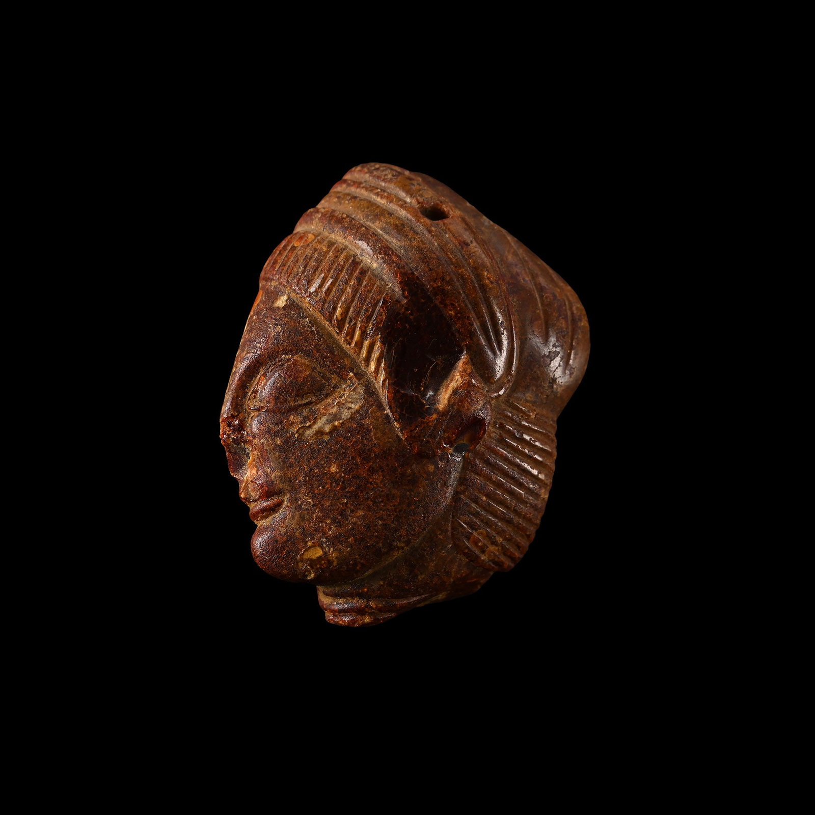 Etruscan Amber Head Pendant: Etruria, 5th century B.C. Carved from translucent red-brown amber as a profile head with stylised Archaic features, large almond eye beneath a defined brow ridge, a softly smiling mouth, and carefully