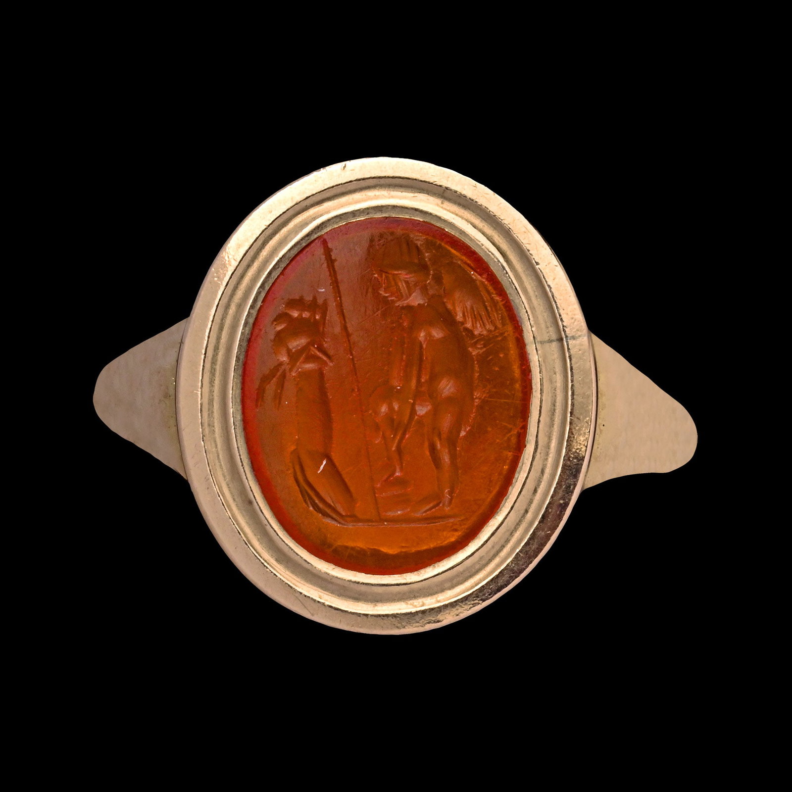 Roman Carnelian Gemstone with Eros Before an Altar in Gold Ring: 1st century A.D. Finely engraved Roman carnelian intaglio showing nude winged Amor in front of a pillar with his weapons, helmet, shield, and spear, putting on a shin guard, standing before an altar, 