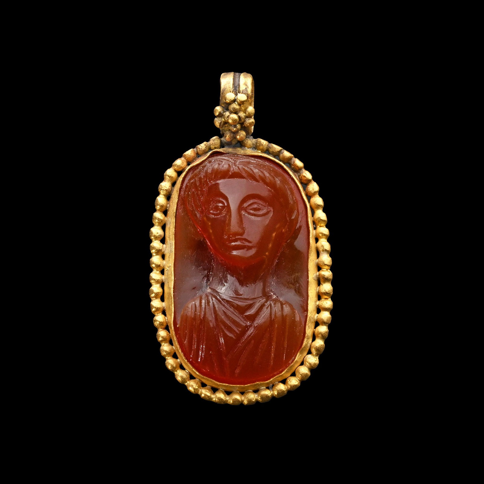 Byzantine Gold Pendant with Carnelian Cameo Bust: Circa 3rd-6th century A.D. Carved bust modelled in the half-round with large lentoid eyes and braided hair to the brow, depicting a disciple, an apostle, or a portrait; gold cell with band of granules