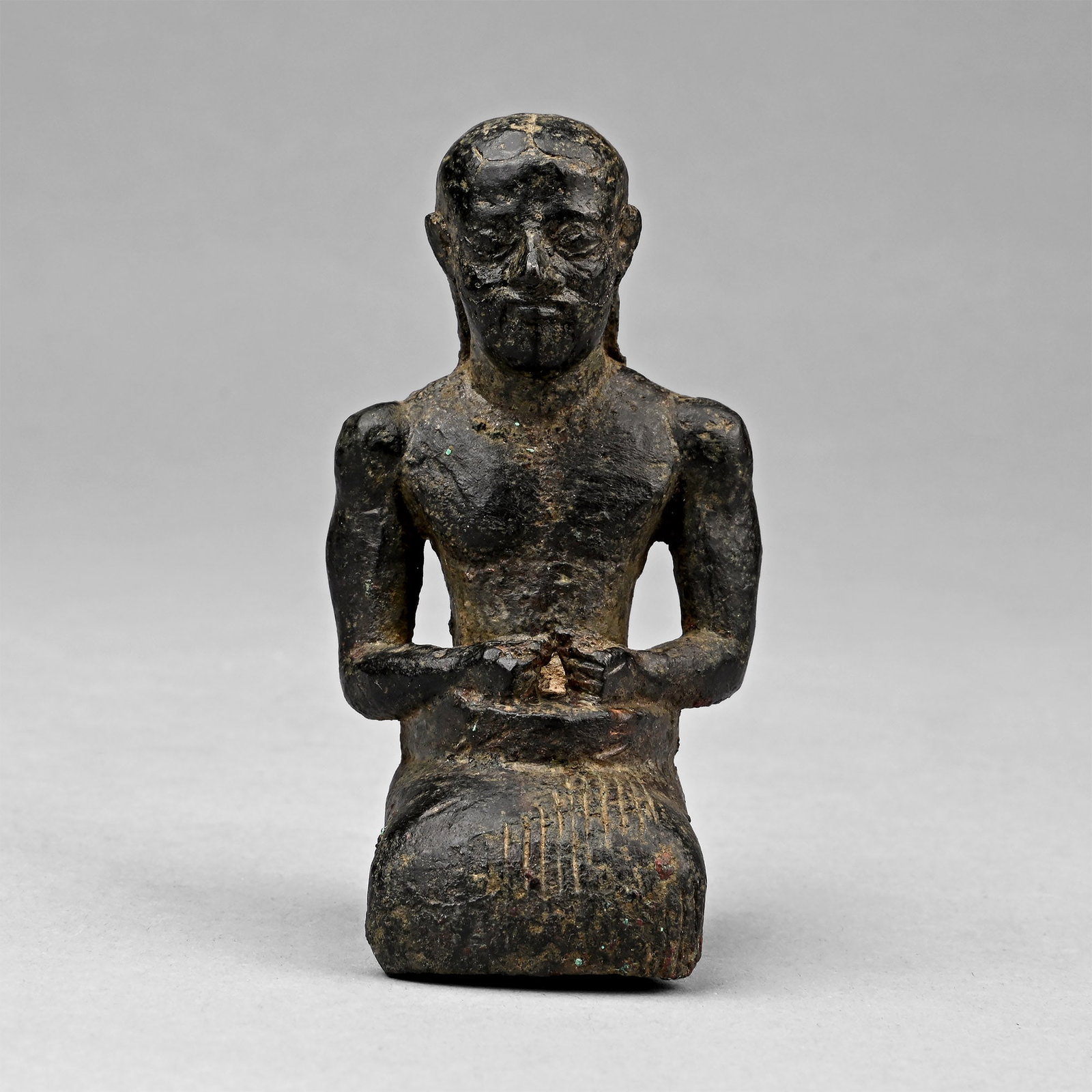 Western Asiatic Bronze Statuette of a Kneeling Priest: Late 3rd millennium B.C. Modelled as a kneeling priest wearing a knee-length kilt, the long hair combed back from the face and extending down the back, arms bent, and fists clenched, resting on the be