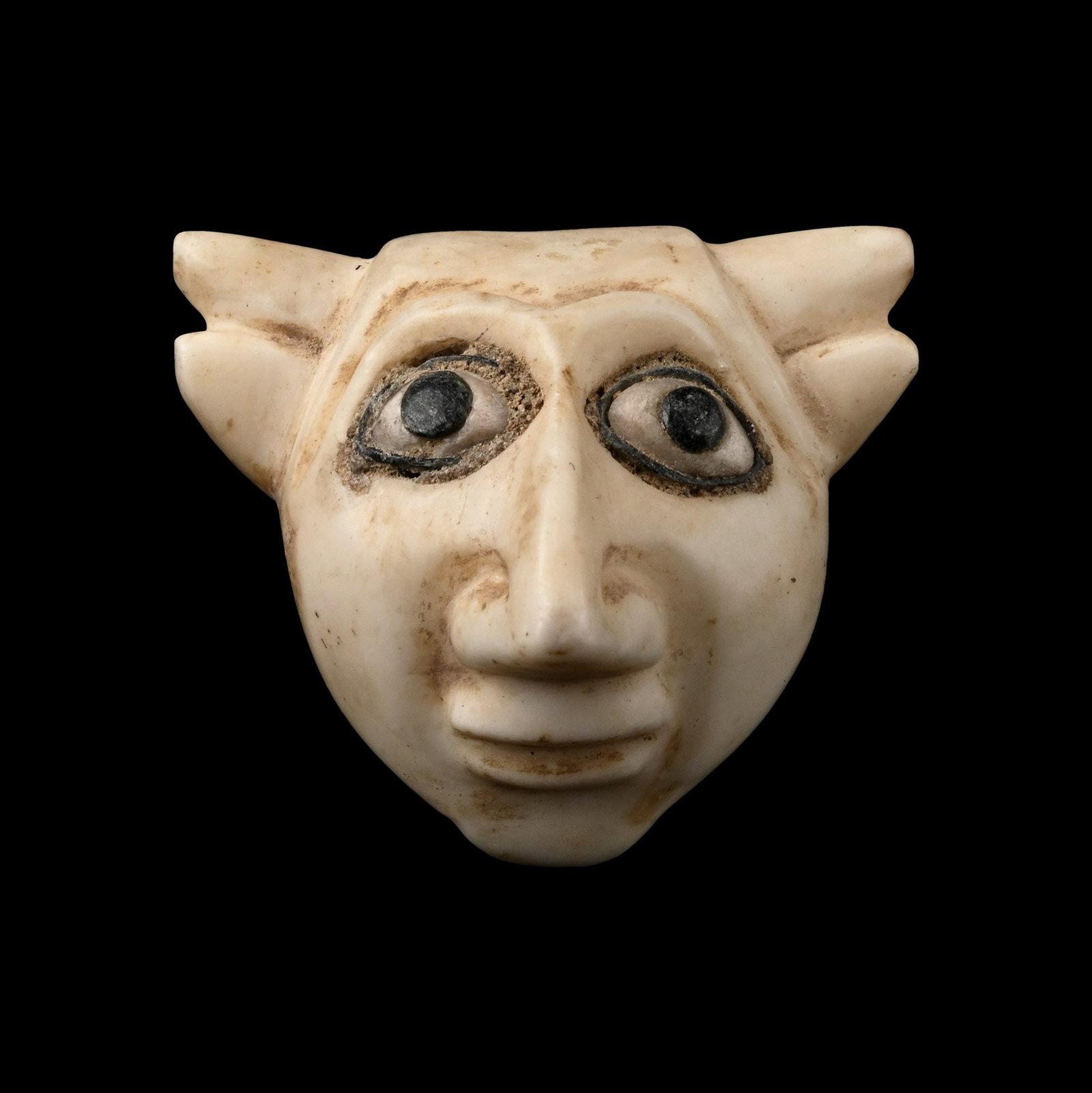 Sumerian Head with Inlaid Eyes: Circa 3000 B.C. D-section carved mask with lateral spurs to the brow, exaggerated aquiline nose and broad mouth with raised lips; applied shell and glass inlay to each eye; pierced at the cheeks for a