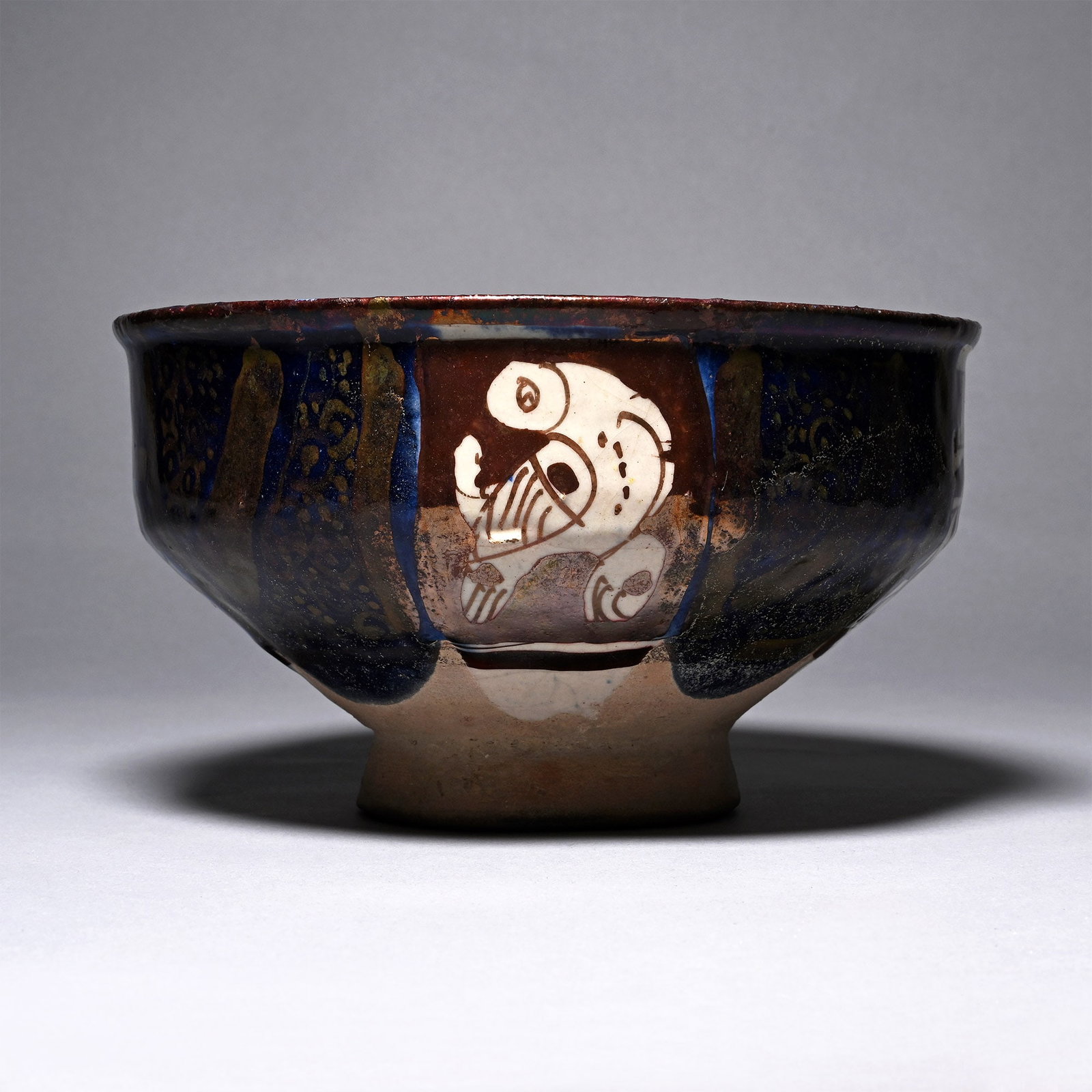 Western Asiatic Cobalt-Blue Footed Ceramic Bowl: 9th century A.D. With a carinated sidewall, rolled and everted rim, shallow foot, partial lustre-glaze to the surface with four trapezoidal panels: two with a reserved regardant bird and two with a tr