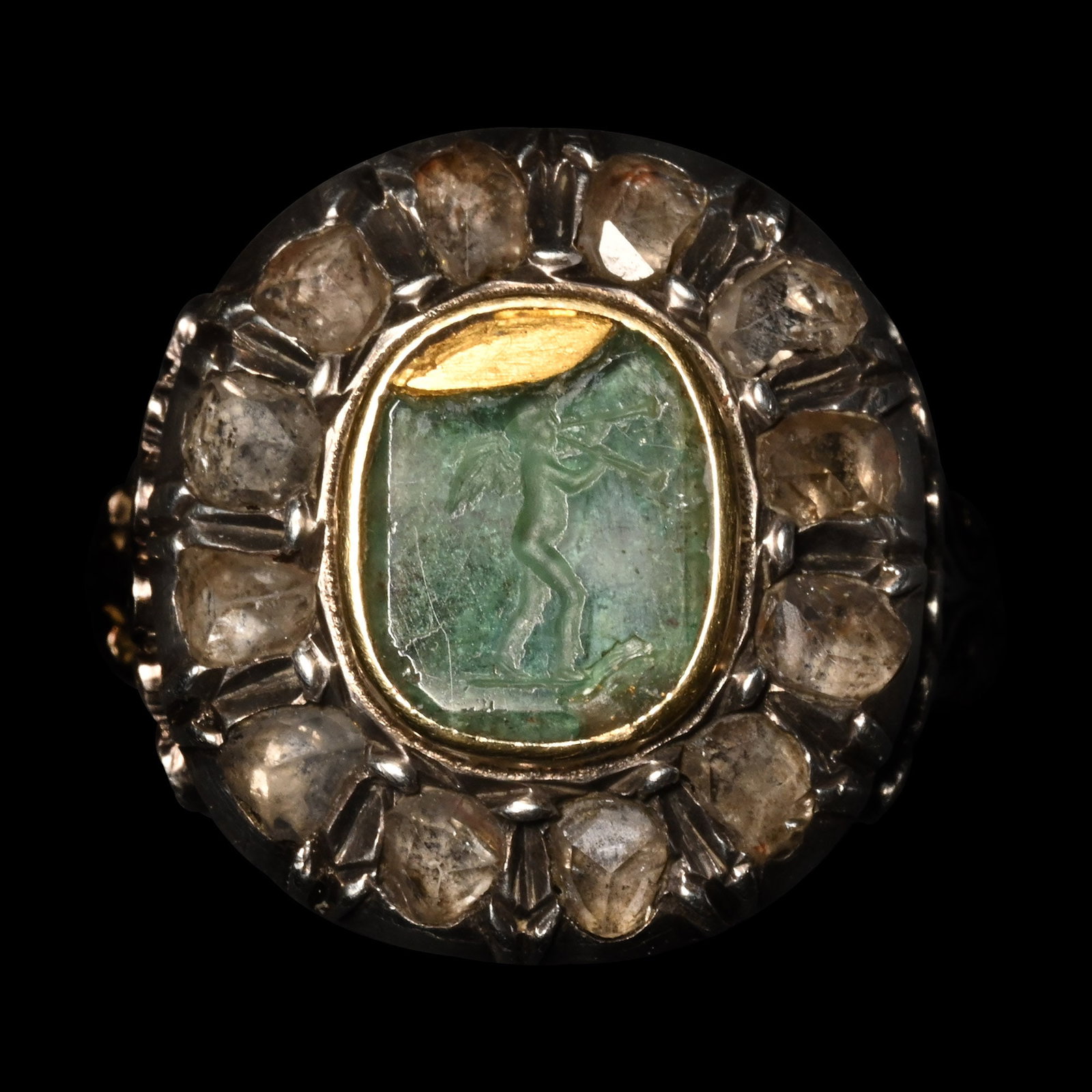 Diamond Locket Ring Set with Emerald Intaglio of Amor, God of Love, Playing Diaulos: Late 19th century A.D. Comprising a segmented hoop with scroll-shaped shoulders, a hinged oval bezel with a secret compartment, and a bezel decorated with twelve cut diamonds around a central green st