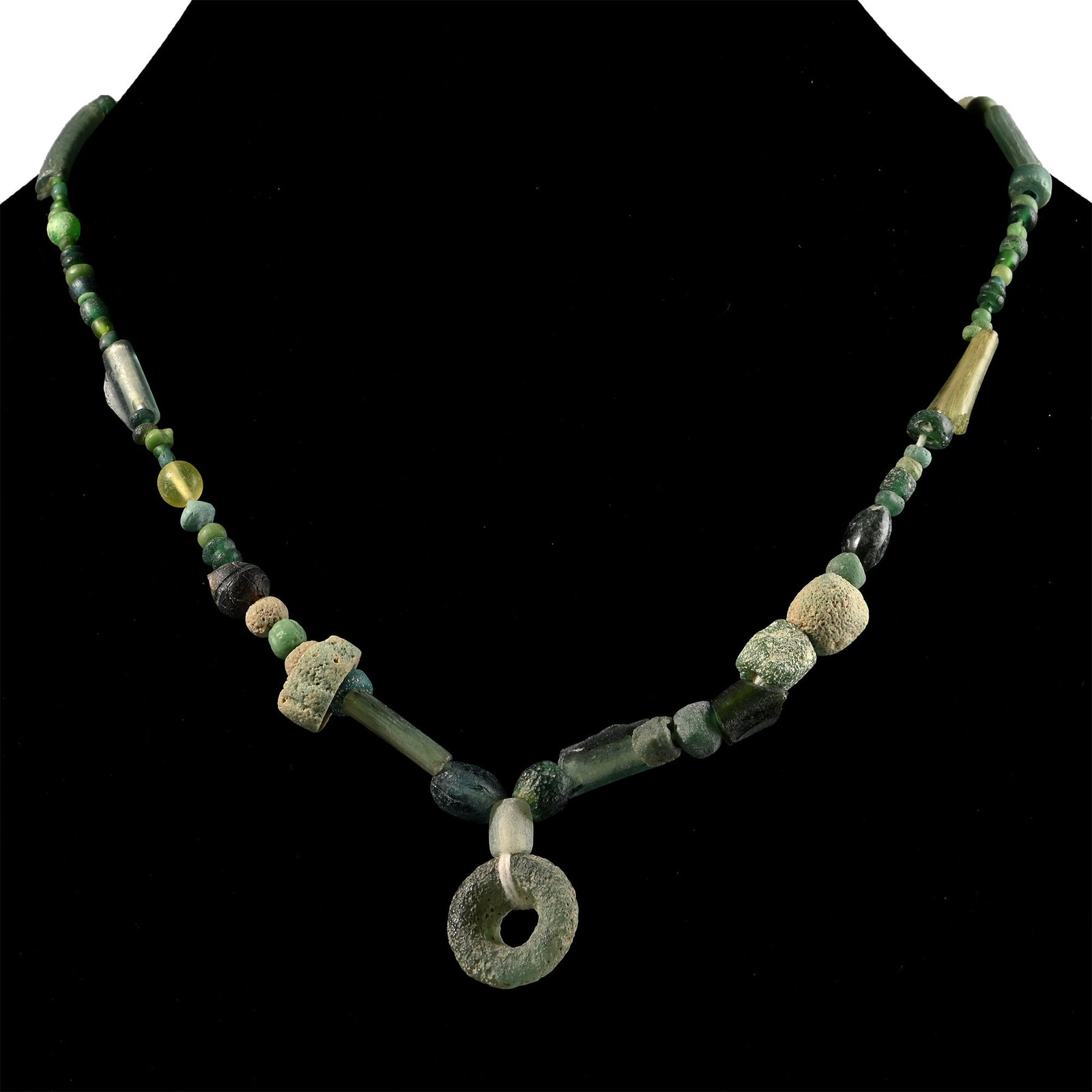 Roman Green Glass Bead Necklace String: 1st-4th century A.D. and later. Composed of mixed beads including oblate, spherical, and other types, the central feature a large annular bead, restrung. 22.8 grams, 41 cm (16 1/8 in.). [No Reserve] A