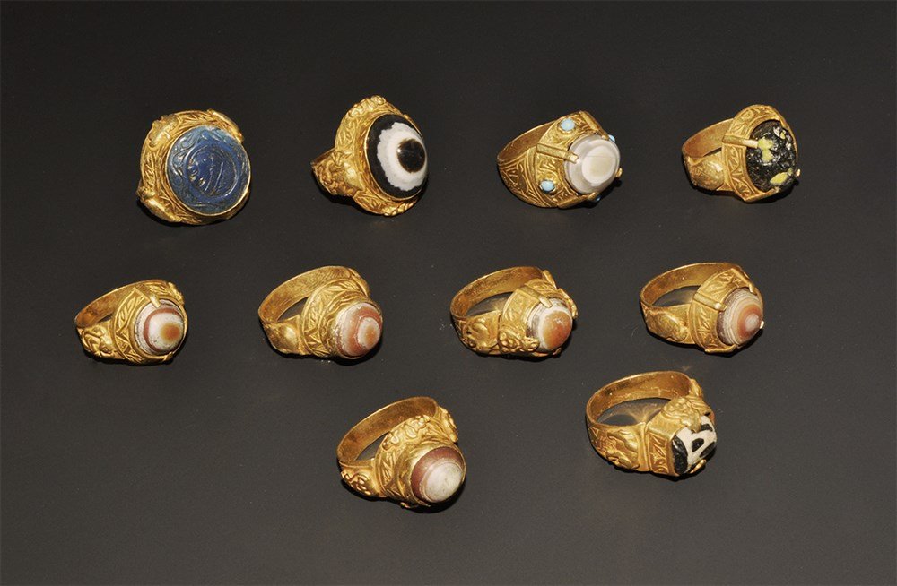 Islamic Style Gilt Metal Ring Group with Intaglio: A mixed group of undated archaistic finger rings comprising: seven with banded agate cabochon; one with blue glass intaglio of a profile male bust; one with blue glass insert with yellow flecks; one w